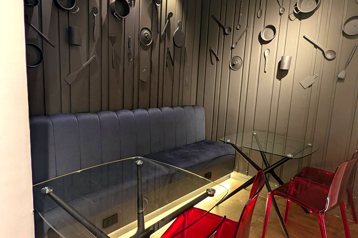 Sala oscura con pared artistica de utensilios de cocina y sillas rojas transparentes